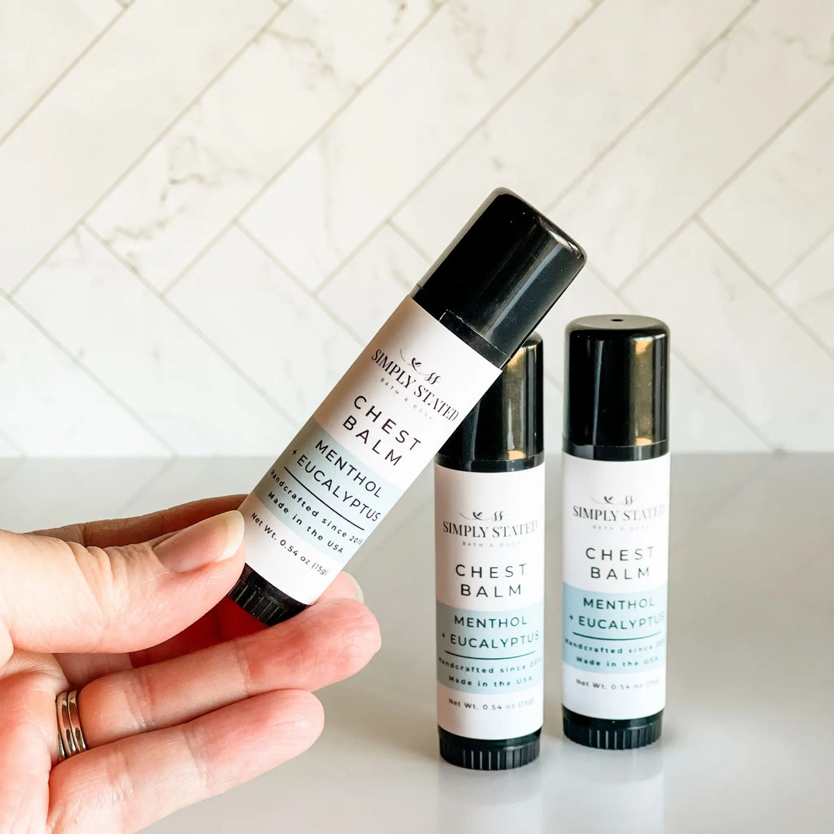 Simply Stated Bath & Body Menthol Eucalyptus Chest Balm stick, one held in hand, two more standing on counter.