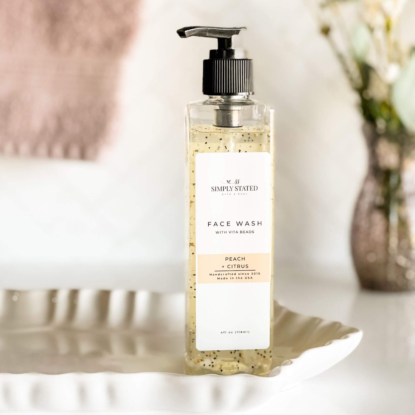 Simply Stated Bath & Body Face Wash with Vita Beads in Peach + Citrus, displayed on a white ceramic tray with soft towel in the background.