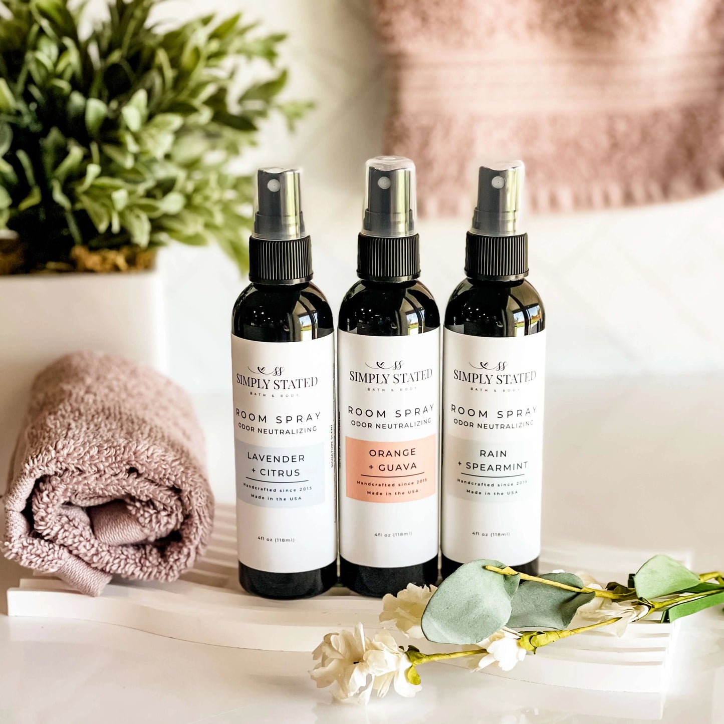 Three room spray bottles from Simply Stated on a surface with a towel and flowers.