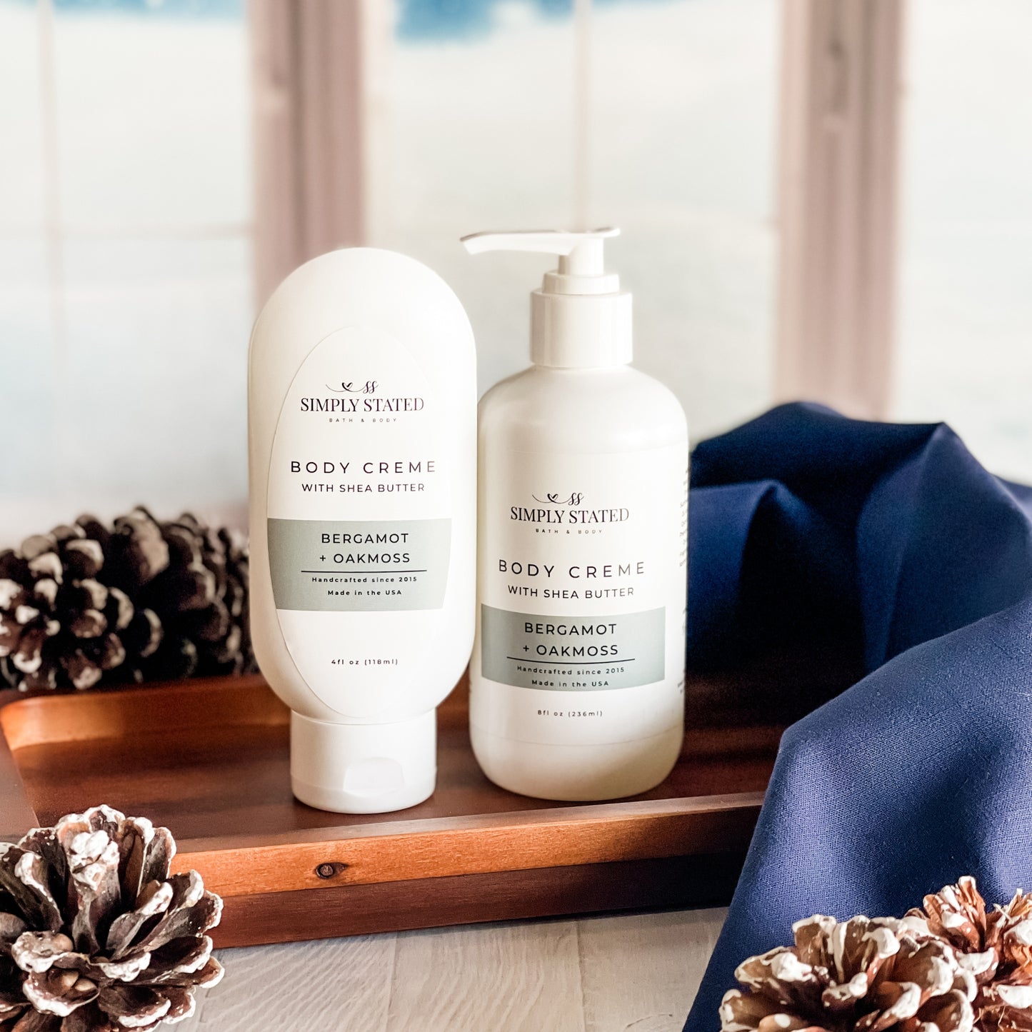 Simply Stated Bath & Body Body Creme with Shea Butter in Bergamot + Oakmoss, tube and pump bottle on wood tray with pinecones