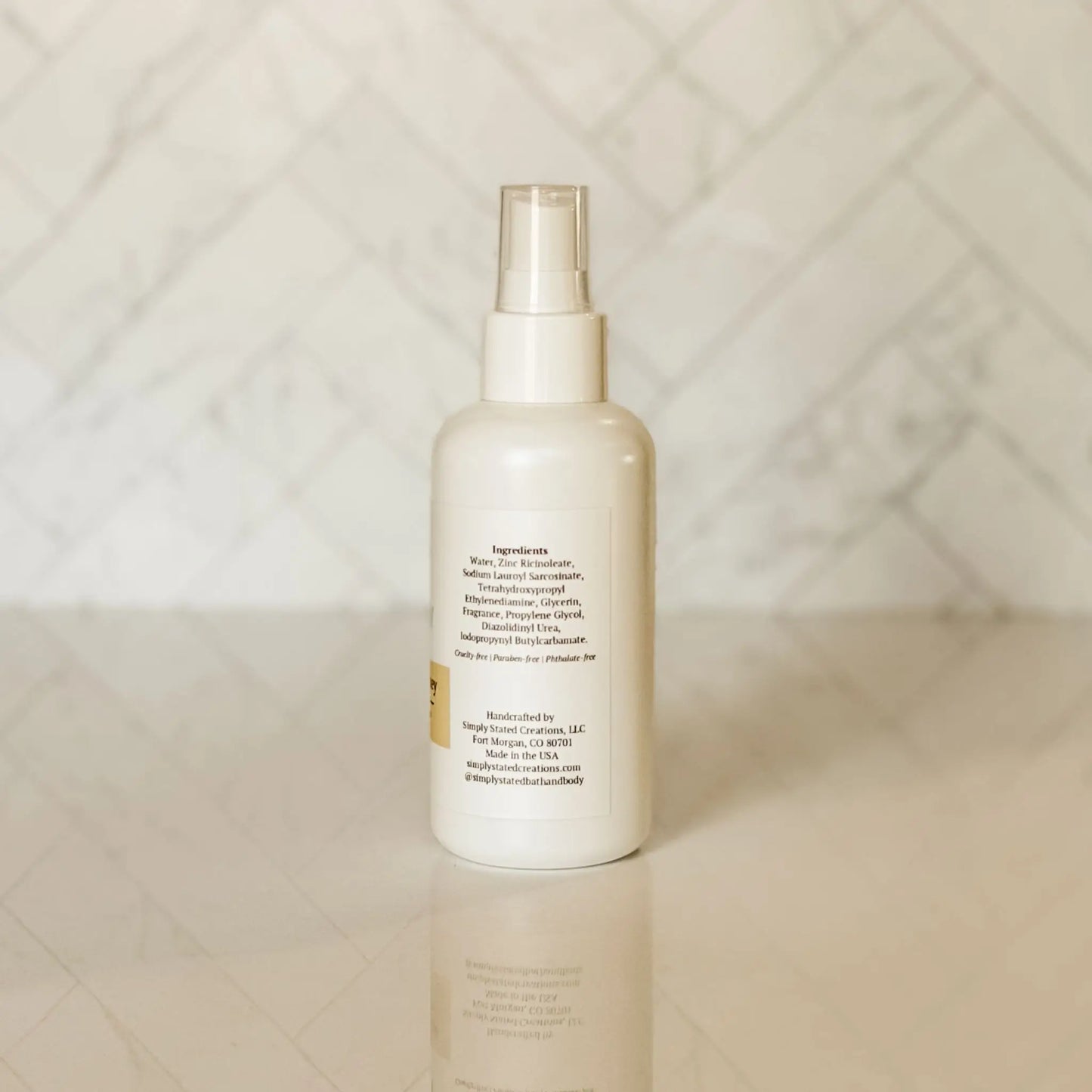 White spray bottle of Simply Stated Bath & Body body mist with ingredients label, set against marble-tiled background