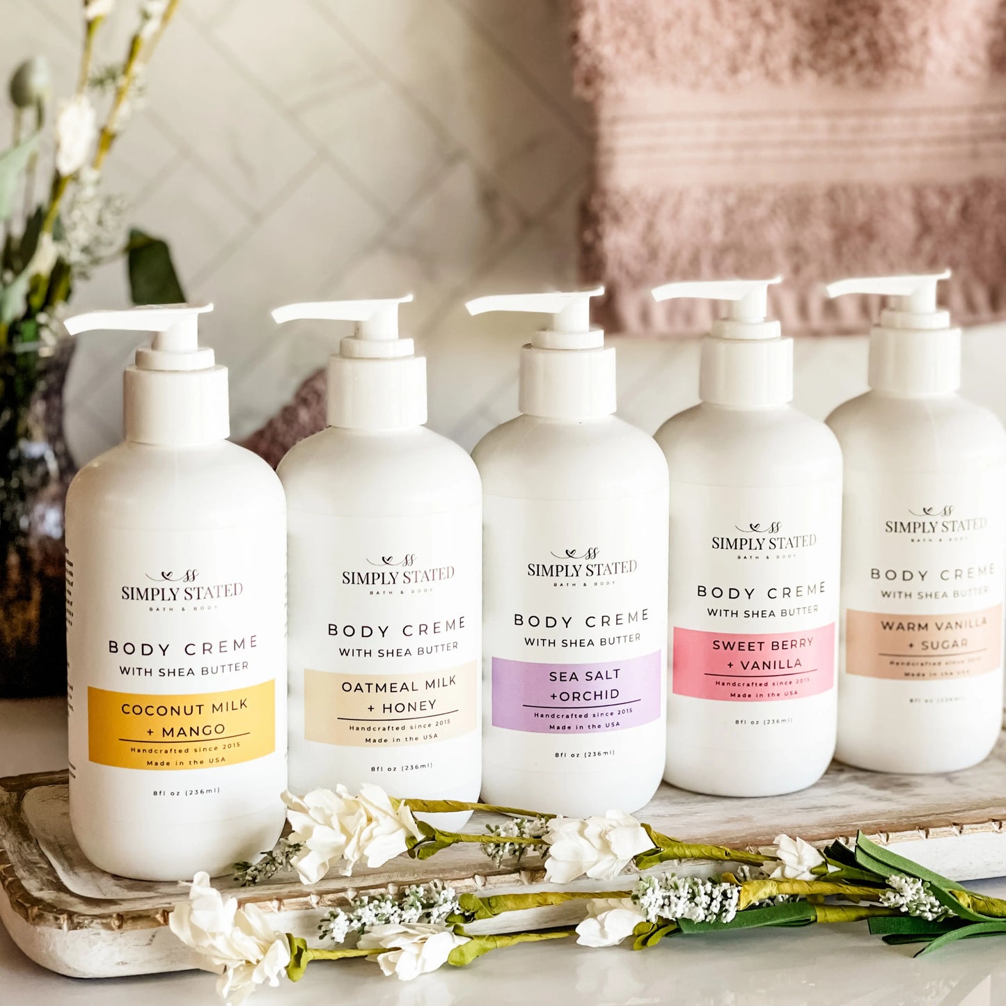 Simply Stated Bath and Body Coconut Milk + Mango Body Creme with four other scents lined up on tray with flowers.