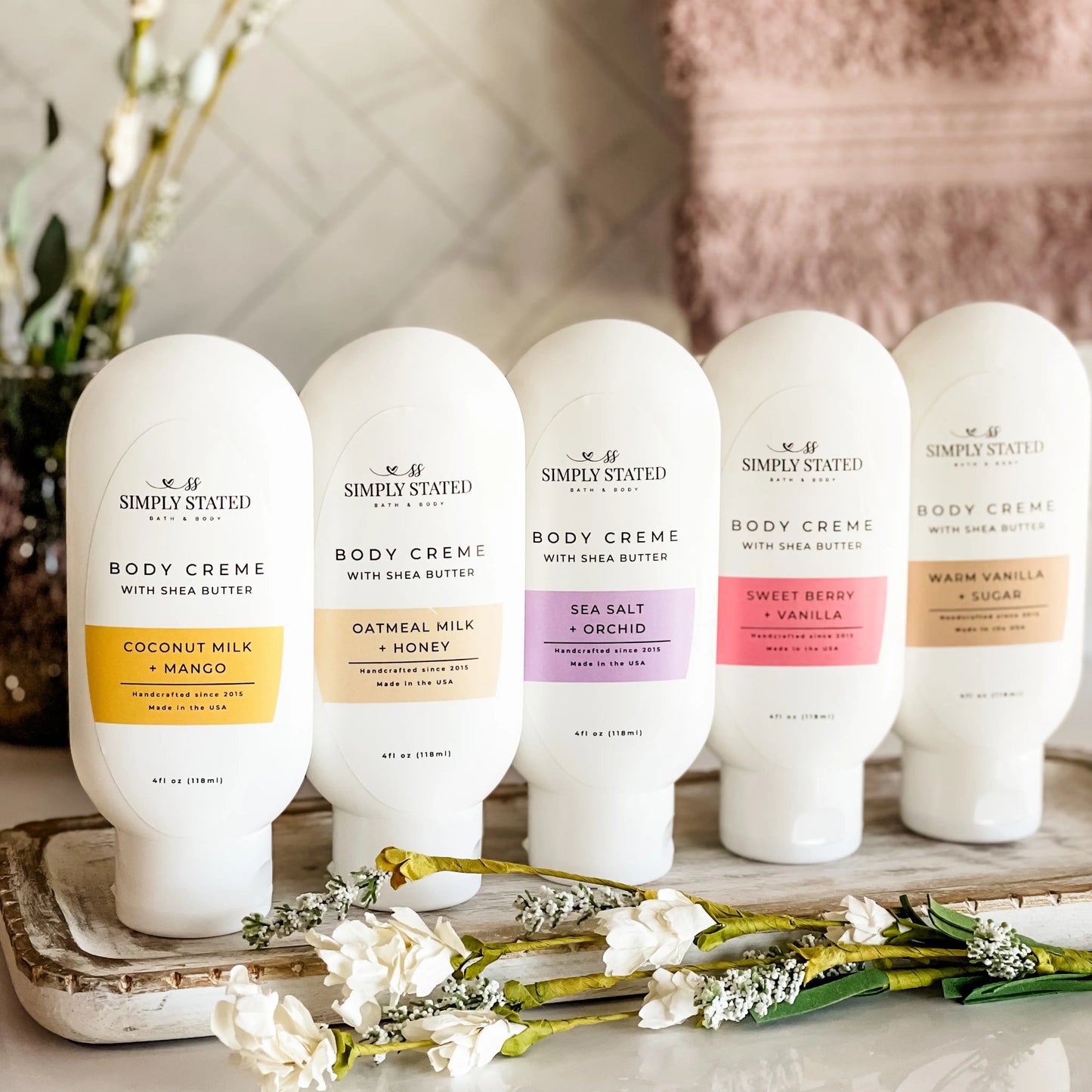 Simply Stated Bath and Body Coconut Milk + Mango Body Creme with four other scents lined up on tray with flowers.