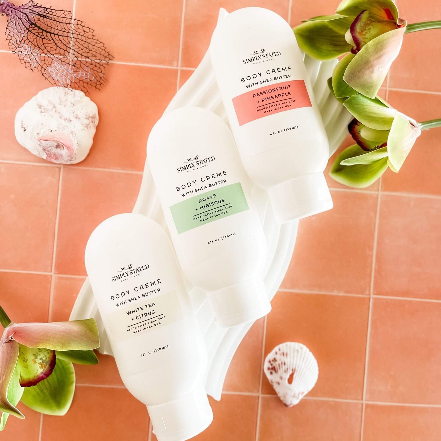 Flatlay of Simply Stated Summer Collection Body Crèmes in Agave + Hibiscus, Passionfruit + Pineapple, and White Tea + Citrus, styled with tropical orchids and coral-toned tiles.