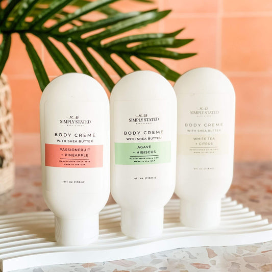Summer Collection Body Crème lineup by Simply Stated Bath & Body, featuring three scents—Passionfruit + Pineapple, Agave + Hibiscus, and White Tea + Citrus—arranged upright on a terrazzo surface with tropical palm decor.