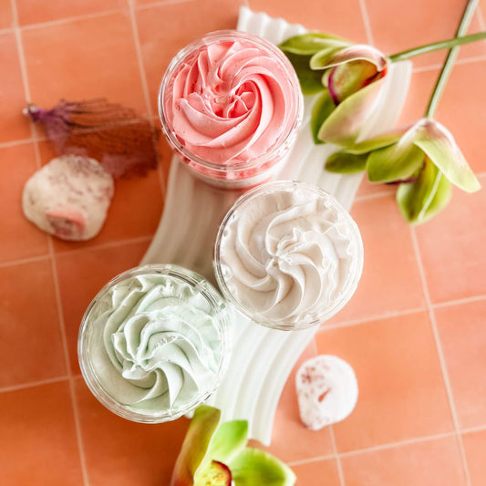 Overhead view of Summer Whipped Soaps in open jars – featuring creamy swirls of Passionfruit + Pineapple, Agave + Hibiscus, and White Tea + Citrus on pink tile with tropical flowers.