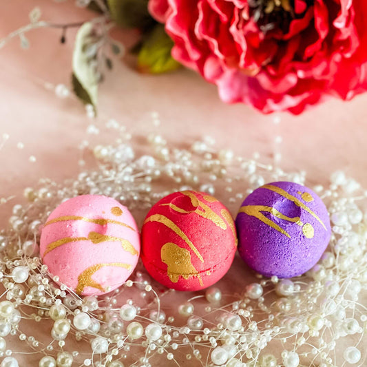 A trio of round bath bombs in pink, red, and purple with gold drizzle accents, displayed on
a soft pink background with pearl decor, representing the Wild Berry Kiss, Raspberry Vanilla Crush, and Strawberry Bubbly fragrances from the Sweet Temptations Collection.
