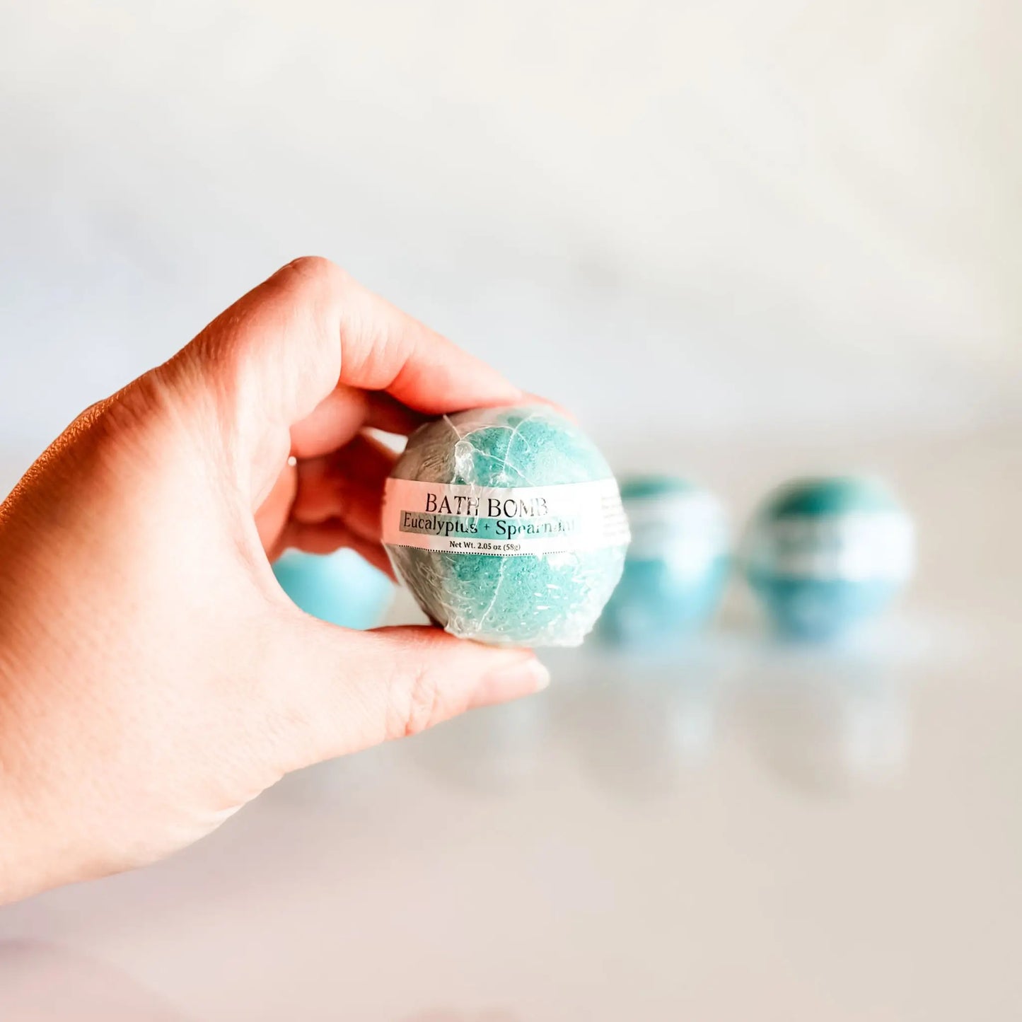 Bath Bomb in Eucalyptus + Spearmint, teal bath bomb held in hand with more bombs blurred in background