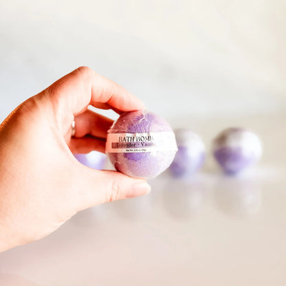 Bath Bomb in Lavender + Vanilla, purple bath bomb held in hand with more bombs blurred in background