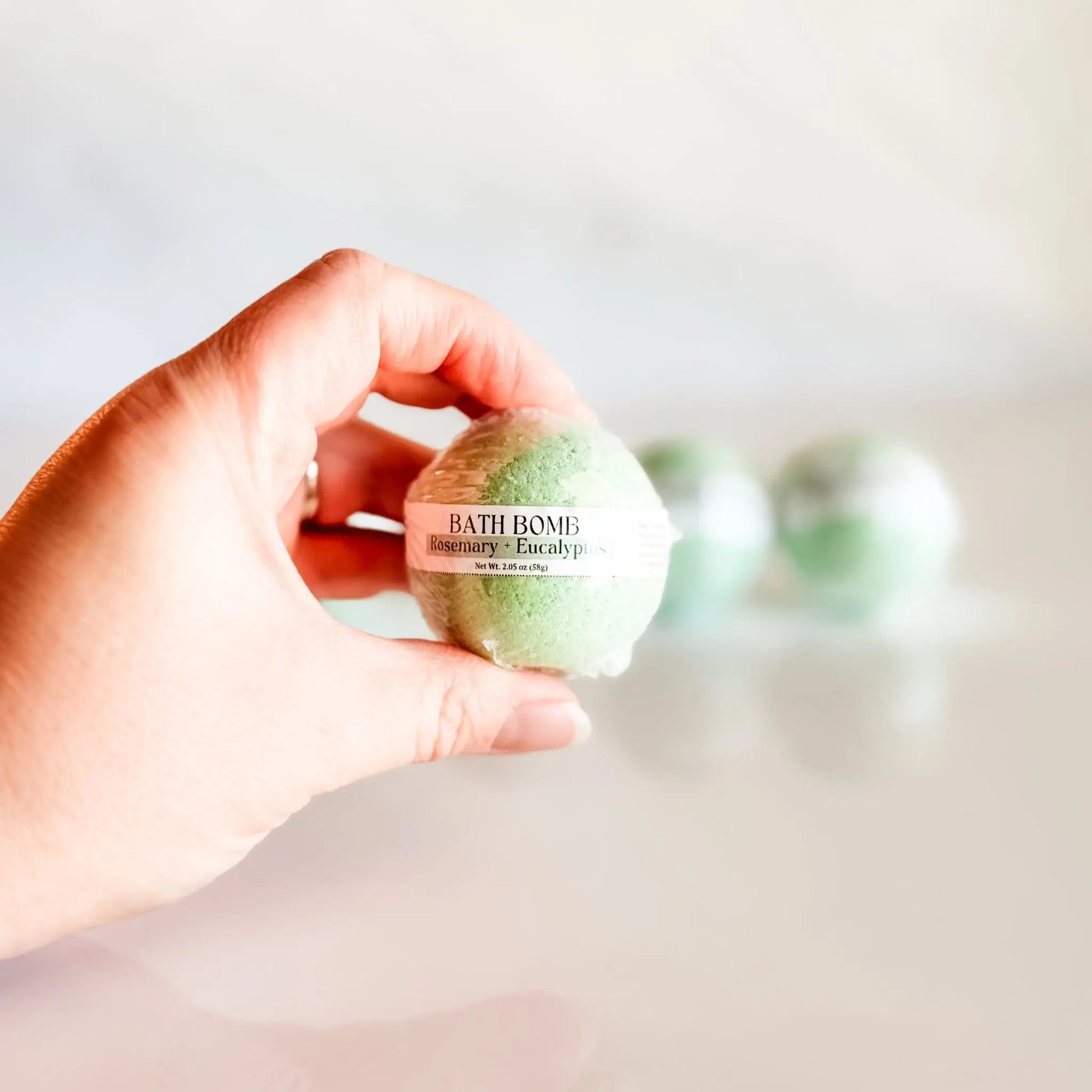 Bath Bomb in Rosemary + Eucalyptus, green bath bomb held in hand with more bombs blurred in background