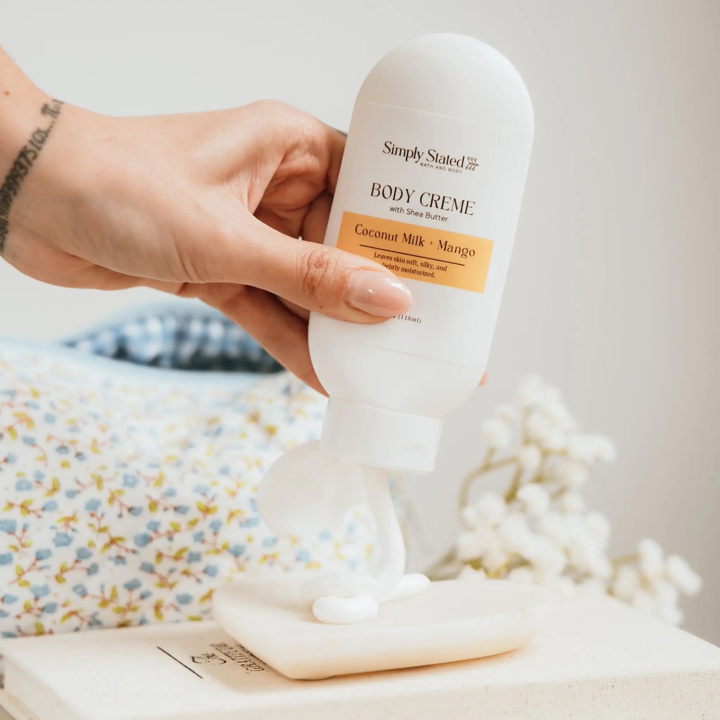 Simply Stated Bath and Body Coconut Milk + Mango Body Creme tube being squeezed onto a soap dish, hand holding tube.