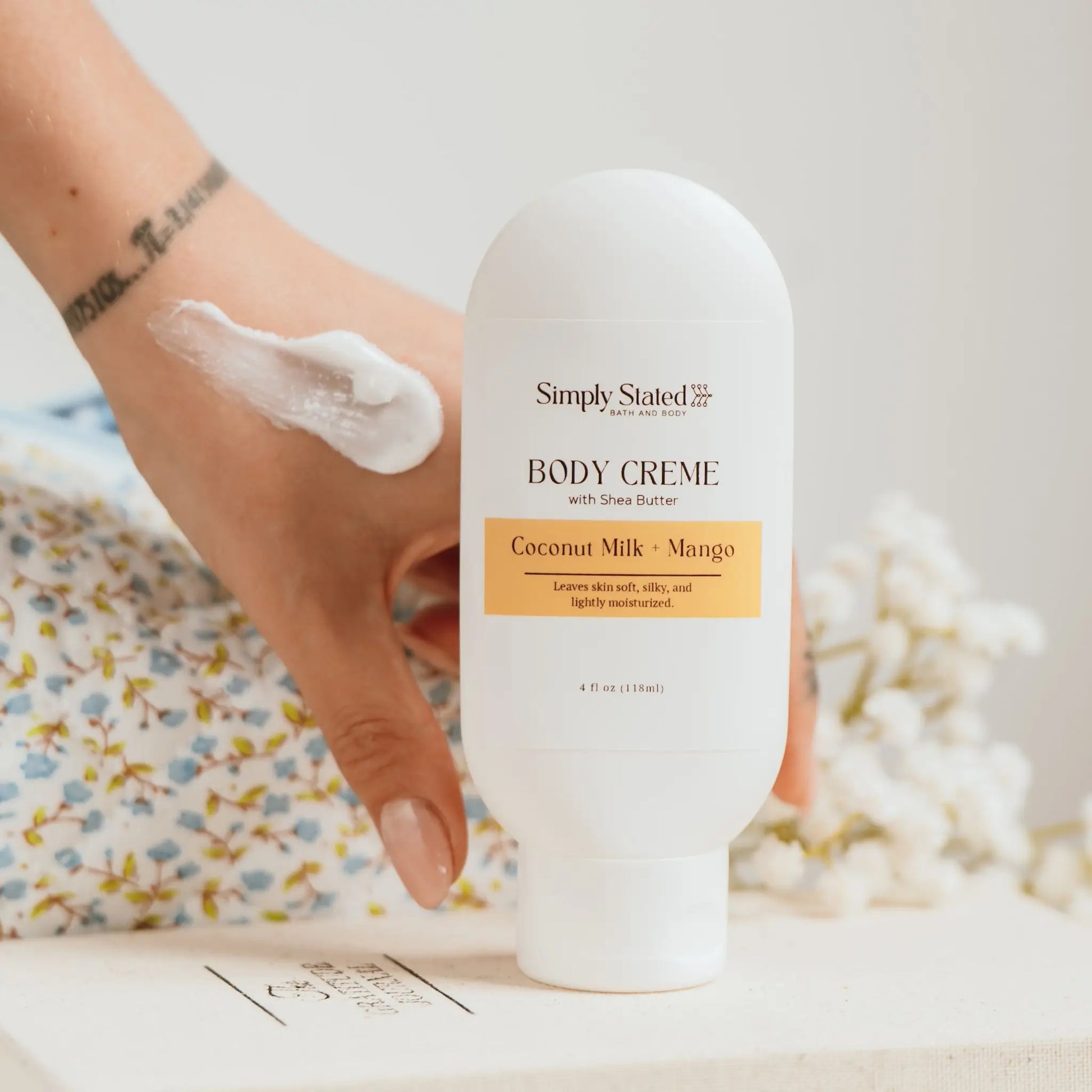 Simply Stated Bath and Body Coconut Milk + Mango Body Creme tube held in hand with cream swatch on wrist, near flowers.