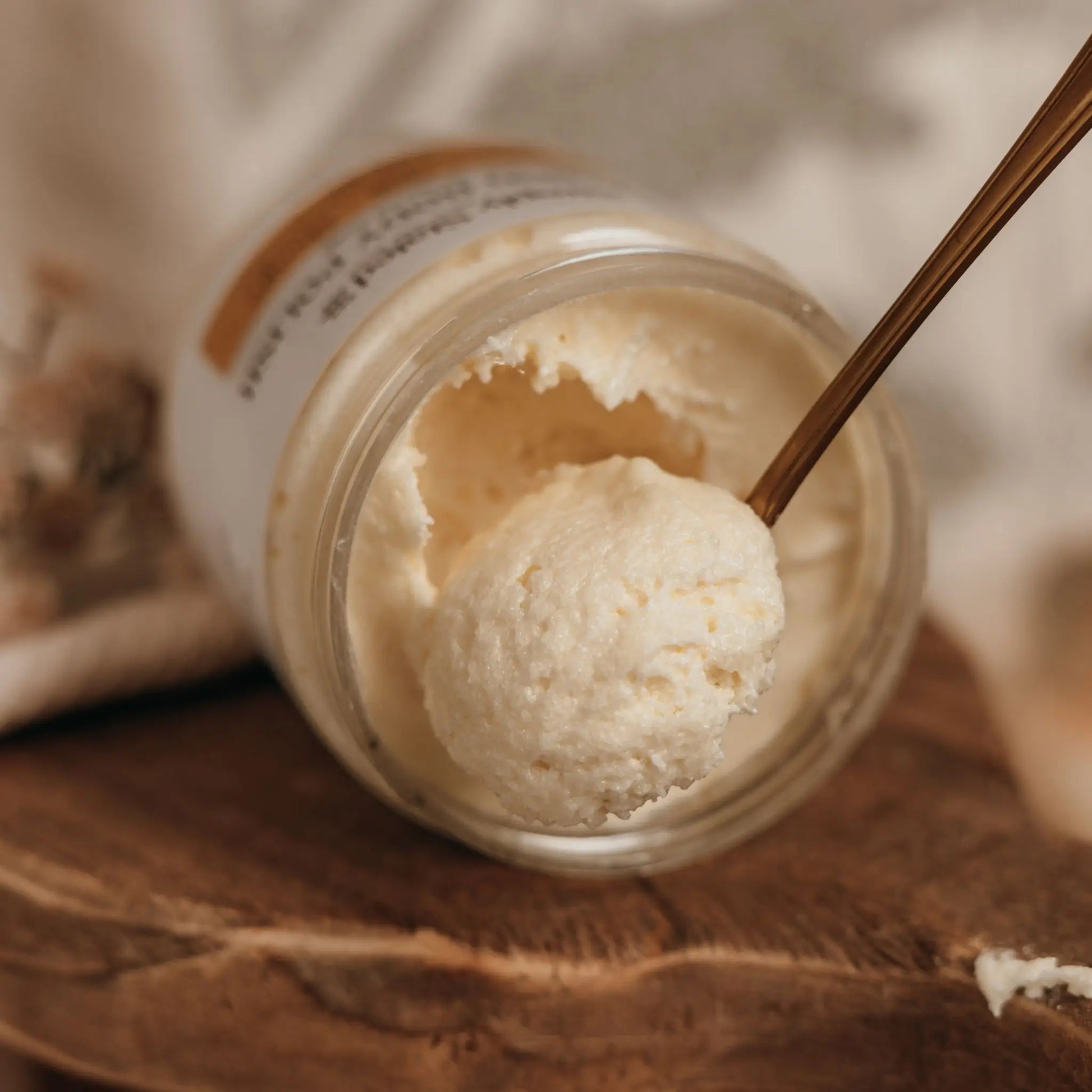 Whipped Body Butter jar open with spoon scooping creamy texture, on wooden surface.