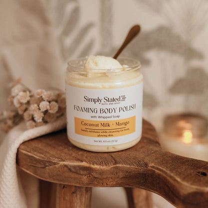 Simply Stated Bath and Body Coconut Milk + Mango Foaming Body Polish jar with spoon on wooden stool, candle nearby.
