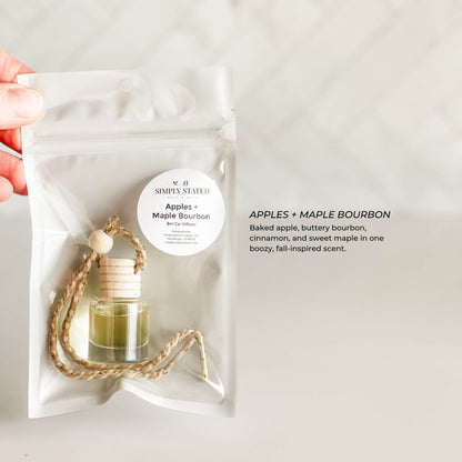 Hand holding a Simply Stated Apple + Maple Bourbon car air freshener packaging on a light gray background.