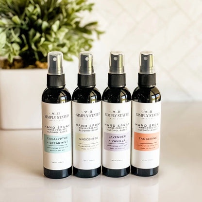 Four bottles of Simply Stated Hand Spray on a light surface with a plant in the background.