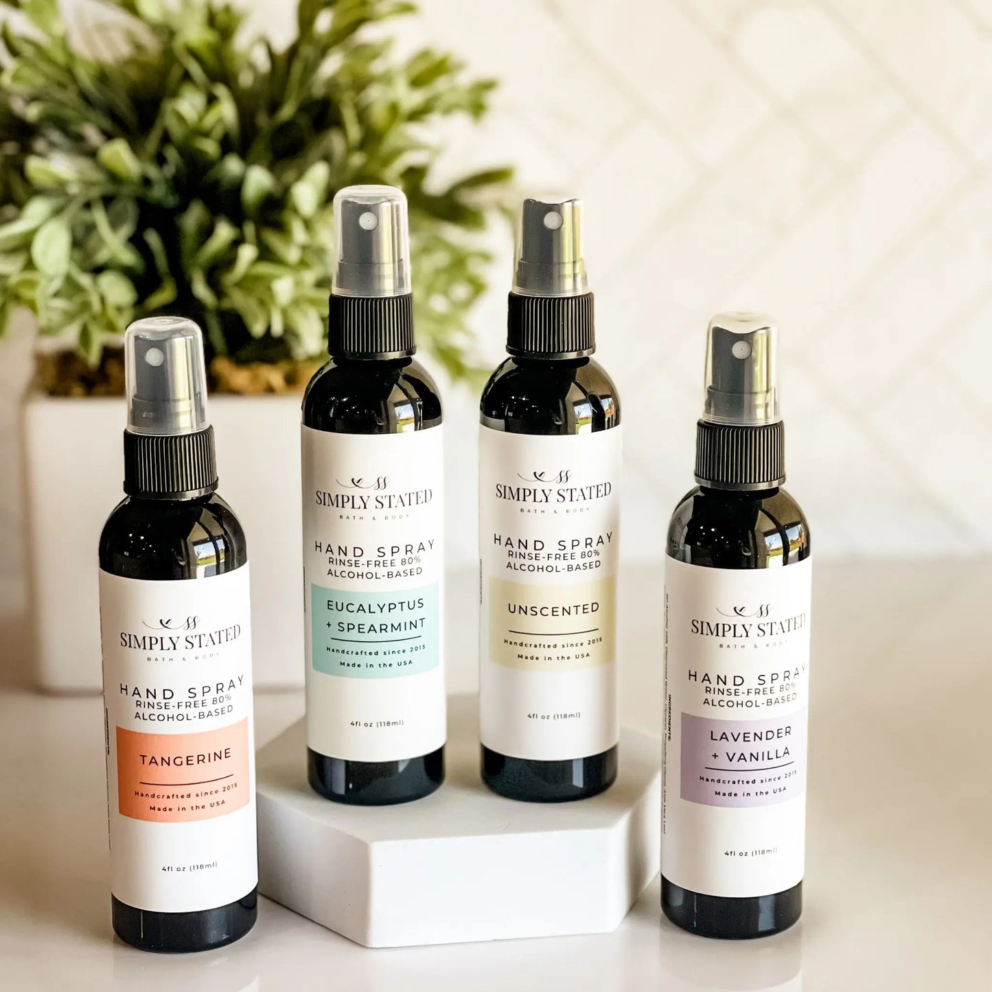 Four bottles of Simply Stated hand spray on a white surface with a plant in the background.