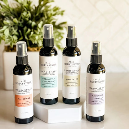 Four bottles of Simply Stated hand spray on a white surface with a plant in the background.