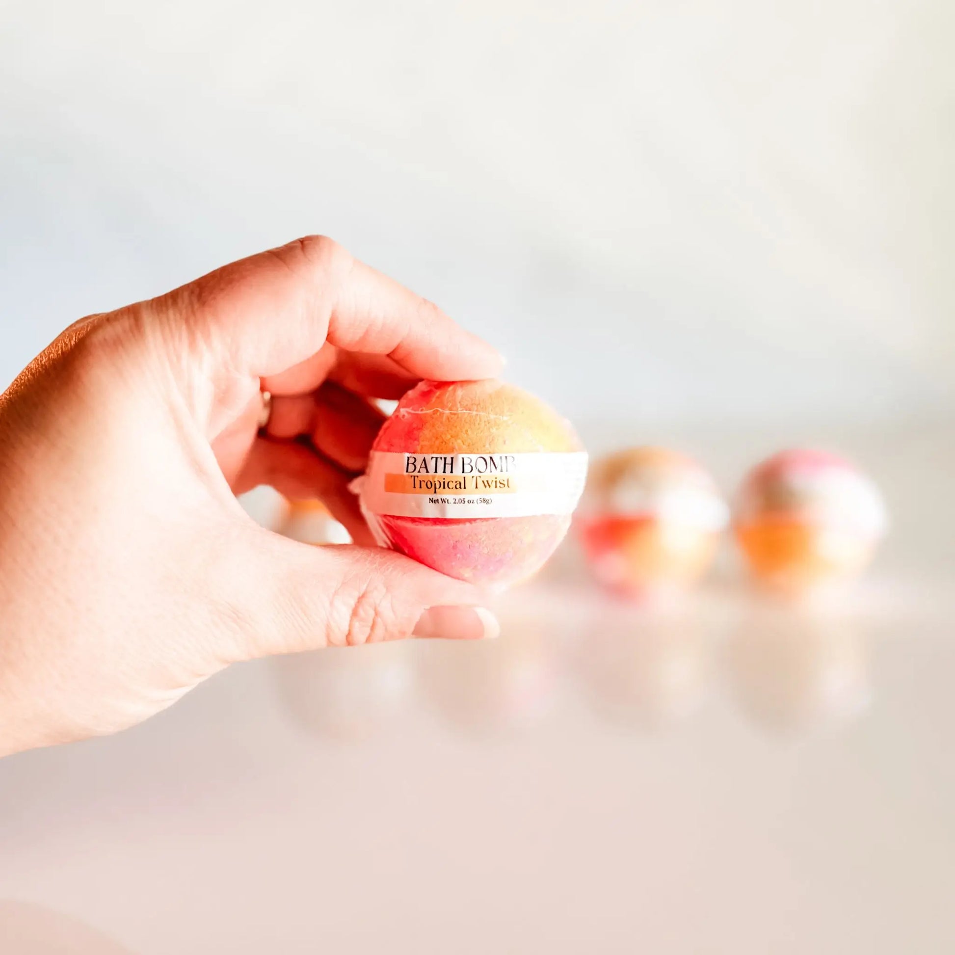 Bath Bomb in Tropical Twist, orange and pink bath bomb held in hand with more bombs blurred in background