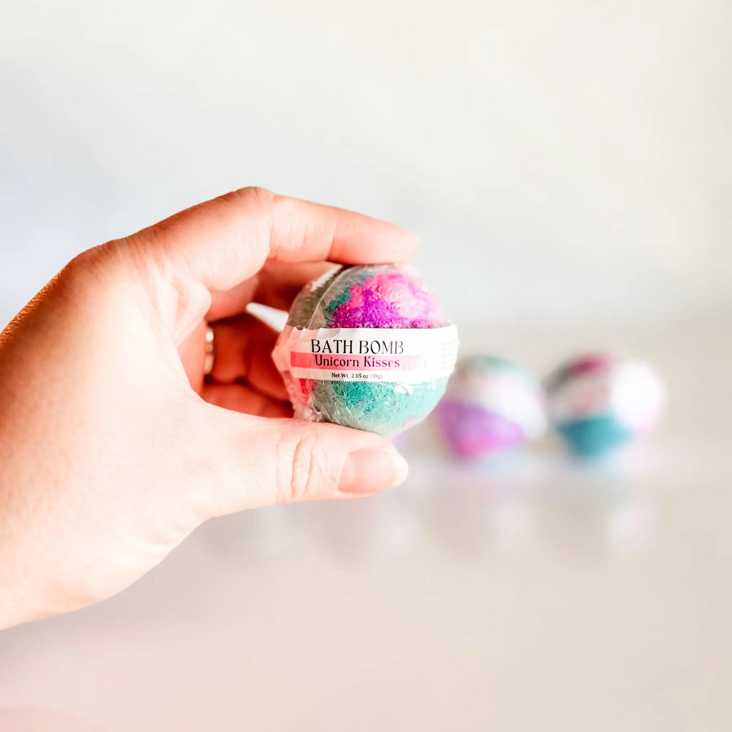 Bath Bomb in Unicorn Kisses, teal and pink bath bomb held in hand with more bombs blurred in background