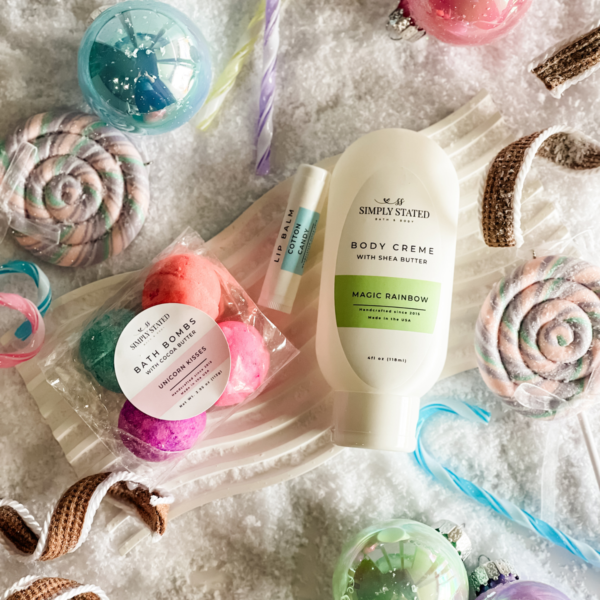 Simply Stated Magic Rainbow Body Creme, Unicorn Kisses Bath Bombs, and Cotton Candy Lip Balm in a pastel candy snow scene.