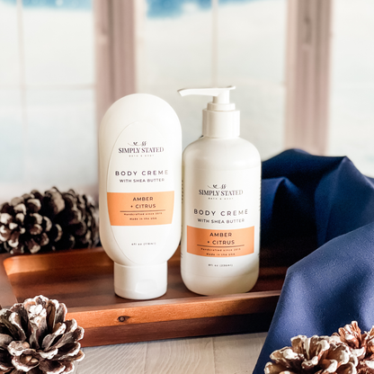Simply Stated Amber + Citrus Body Creme with shea butter, tube and pump sizes on a wood tray with pinecones and navy linen.