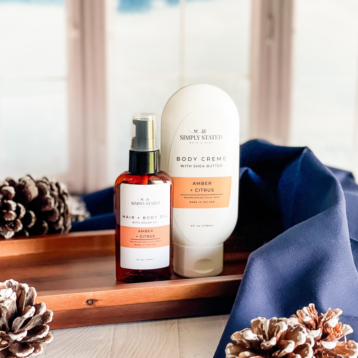 Simply Stated amber citrus hair and body oil with argan oil and shea butter body creme beside pinecones and navy linen