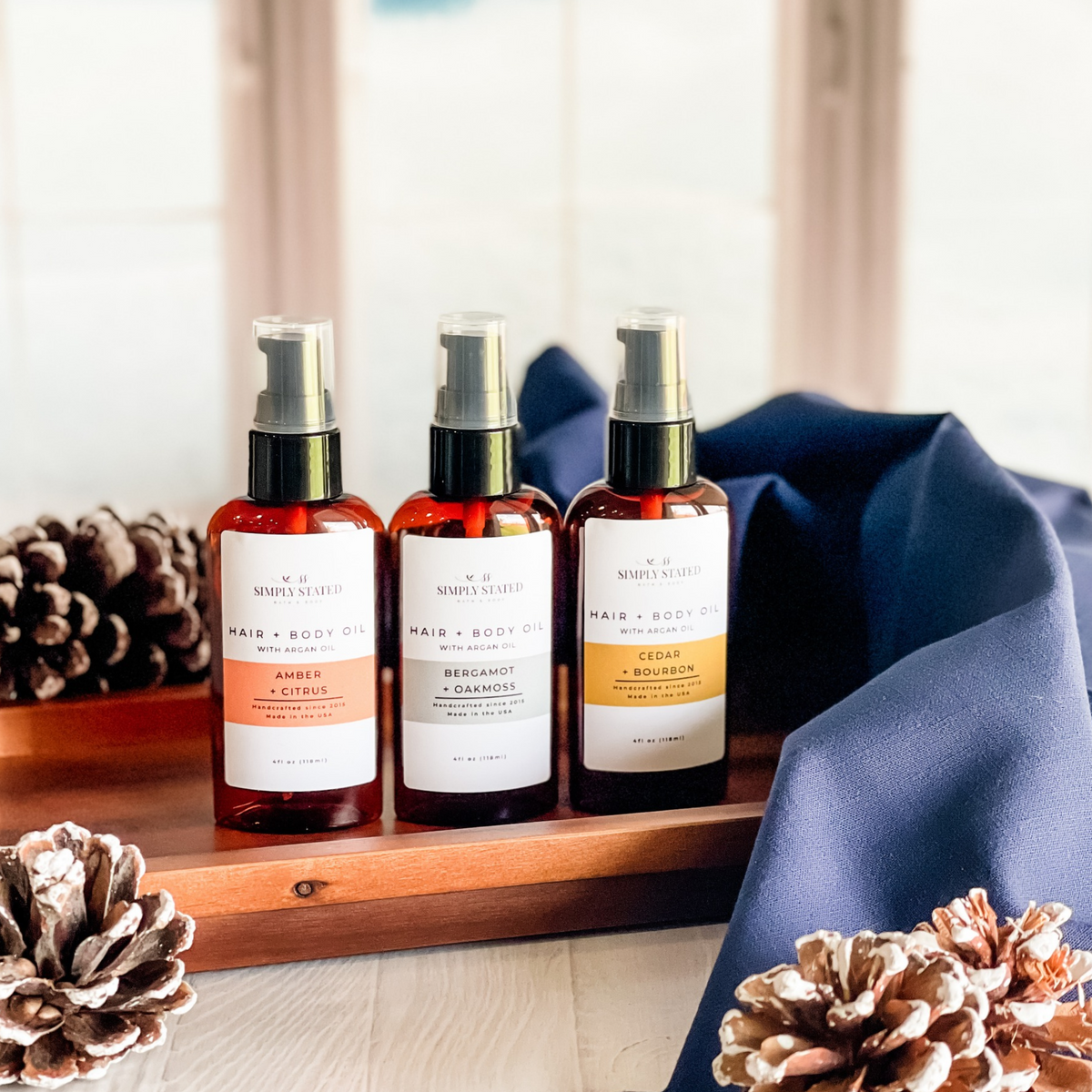 Simply Stated hair and body oil with argan oil in three scents beside frosted pinecones and a navy cloth