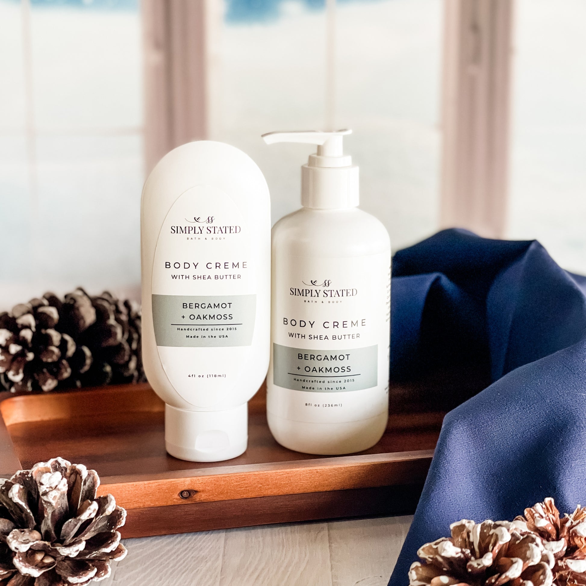 Two bottles of Simply Stated Body Creme on a wooden tray with decorative pinecones.