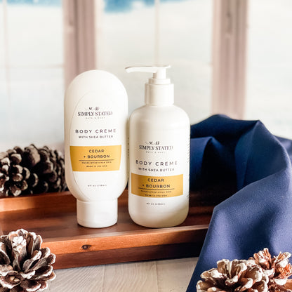 Two bottles of 'Simply Stated' body crème on a wooden tray with decorative pinecones.