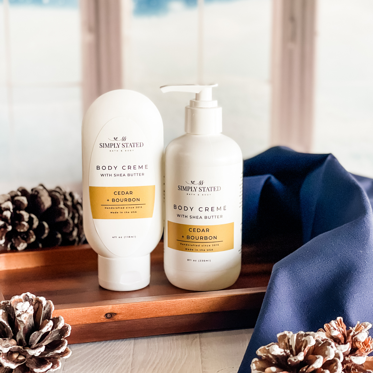 Simply Stated Cedar + Bourbon Body Creme with shea butter, tube and pump sizes on a wood tray with pinecones and navy linen.