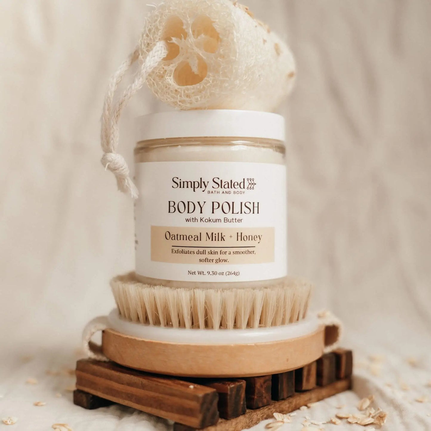 Simply Stated Bath and Body Oatmeal Milk + Honey Body Polish jar on wooden brush with loofah, oats scattered around.