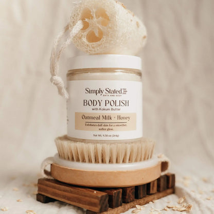 Simply Stated Bath and Body Oatmeal Milk + Honey Body Polish jar on wooden brush with loofah, oats scattered around.