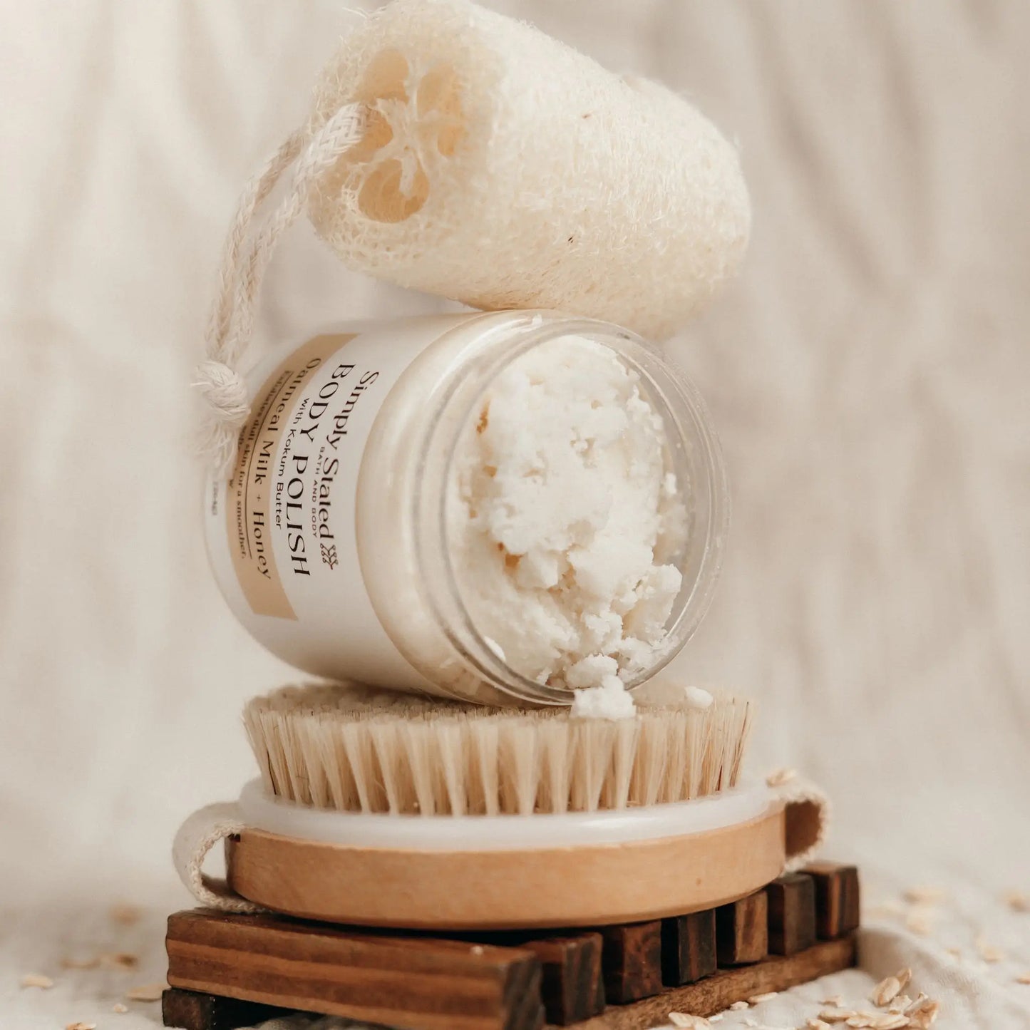 Simply Stated Bath and Body Oatmeal Milk + Honey Body Polish tilted jar showing scrub texture, on brush with loofah.