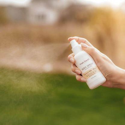 Oatmeal Milk + Honey Body Spray being sprayed outdoors, hand holding bottle against green background.