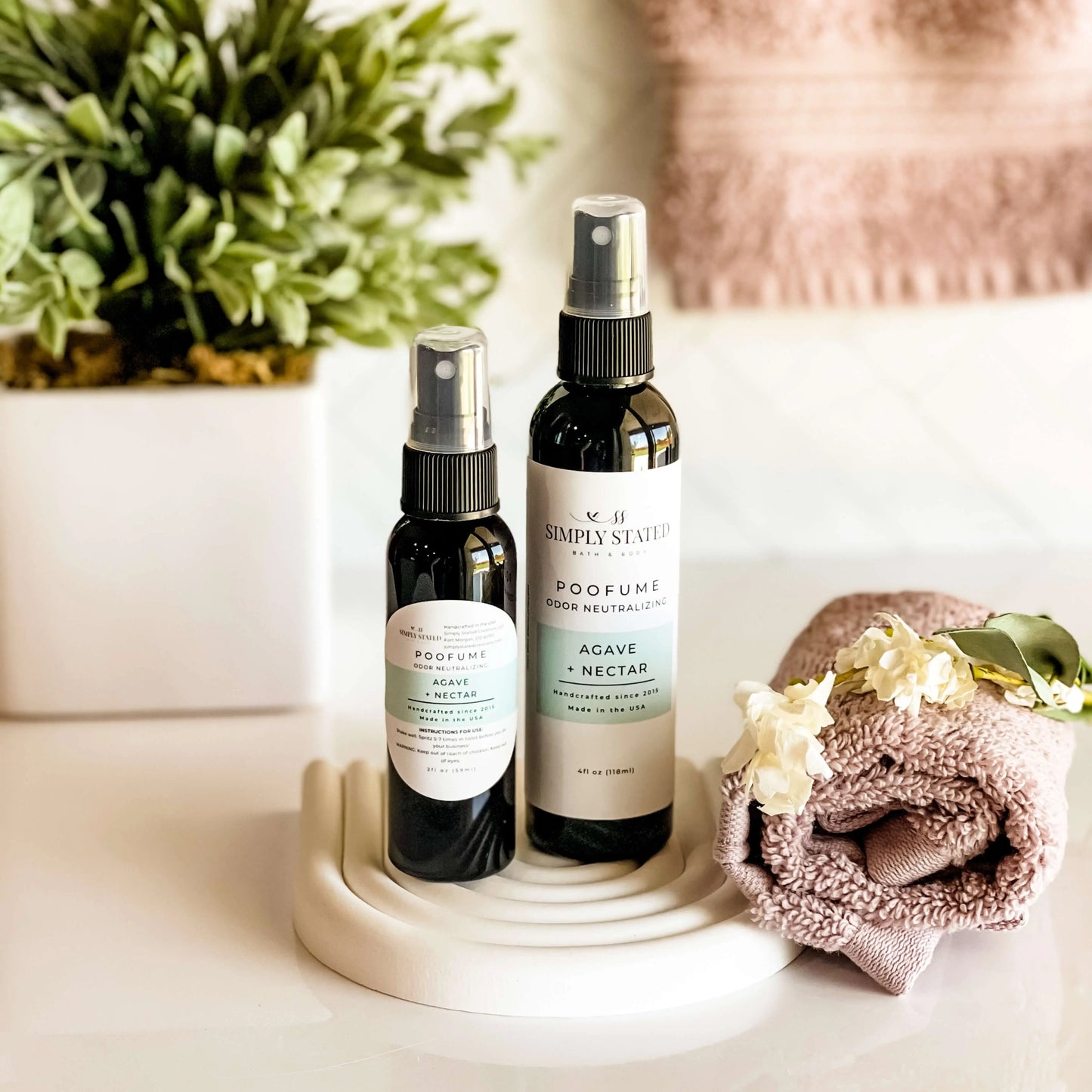 Two spray bottles on a white surface with a plant and towel in the background