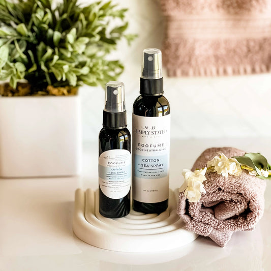 Two spray bottles labeled 'Cotton + Sea Spray' and 'Simply Stated' on a white surface with a towel and plant in the background.