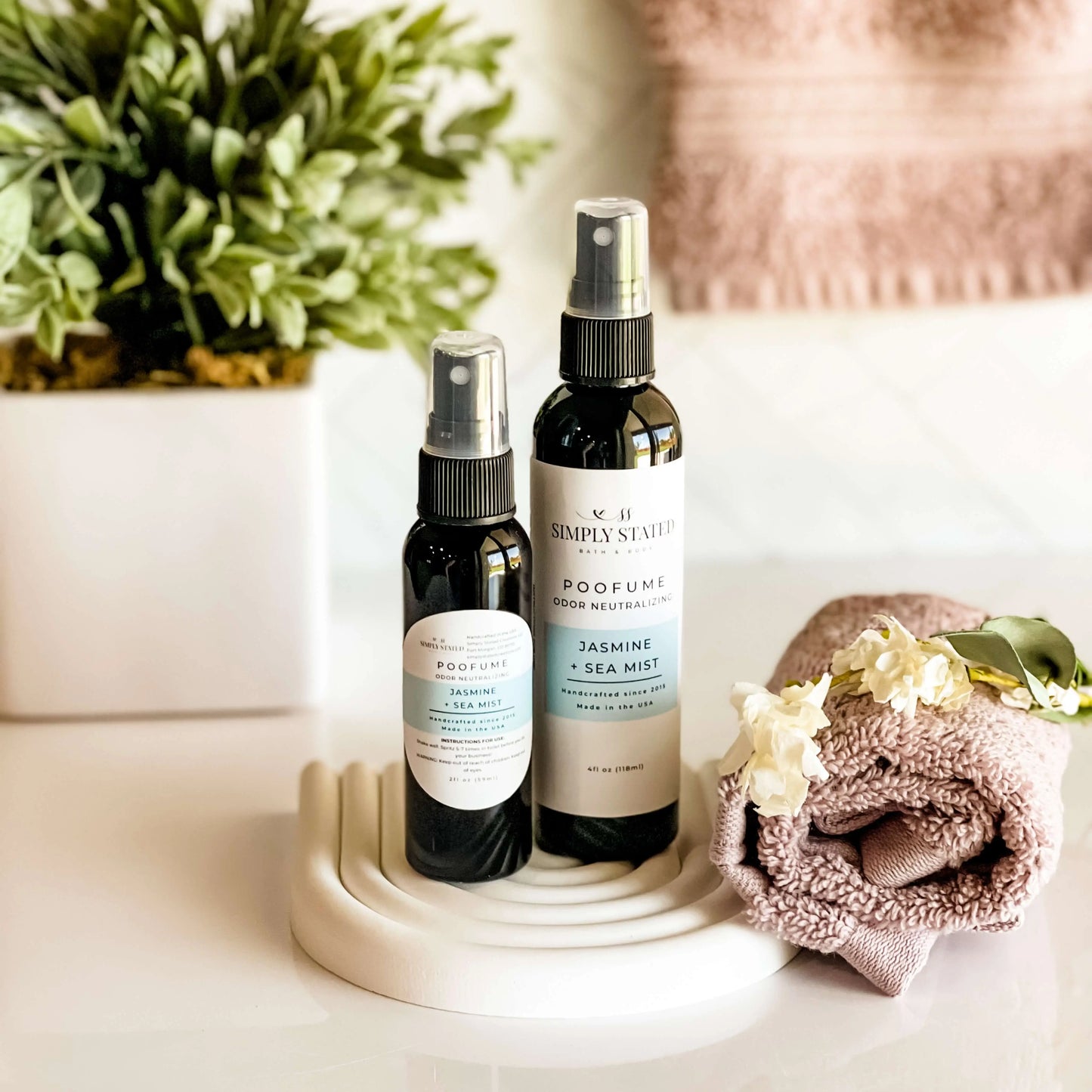 Two spray bottles on a white surface with a plant and towel in the background