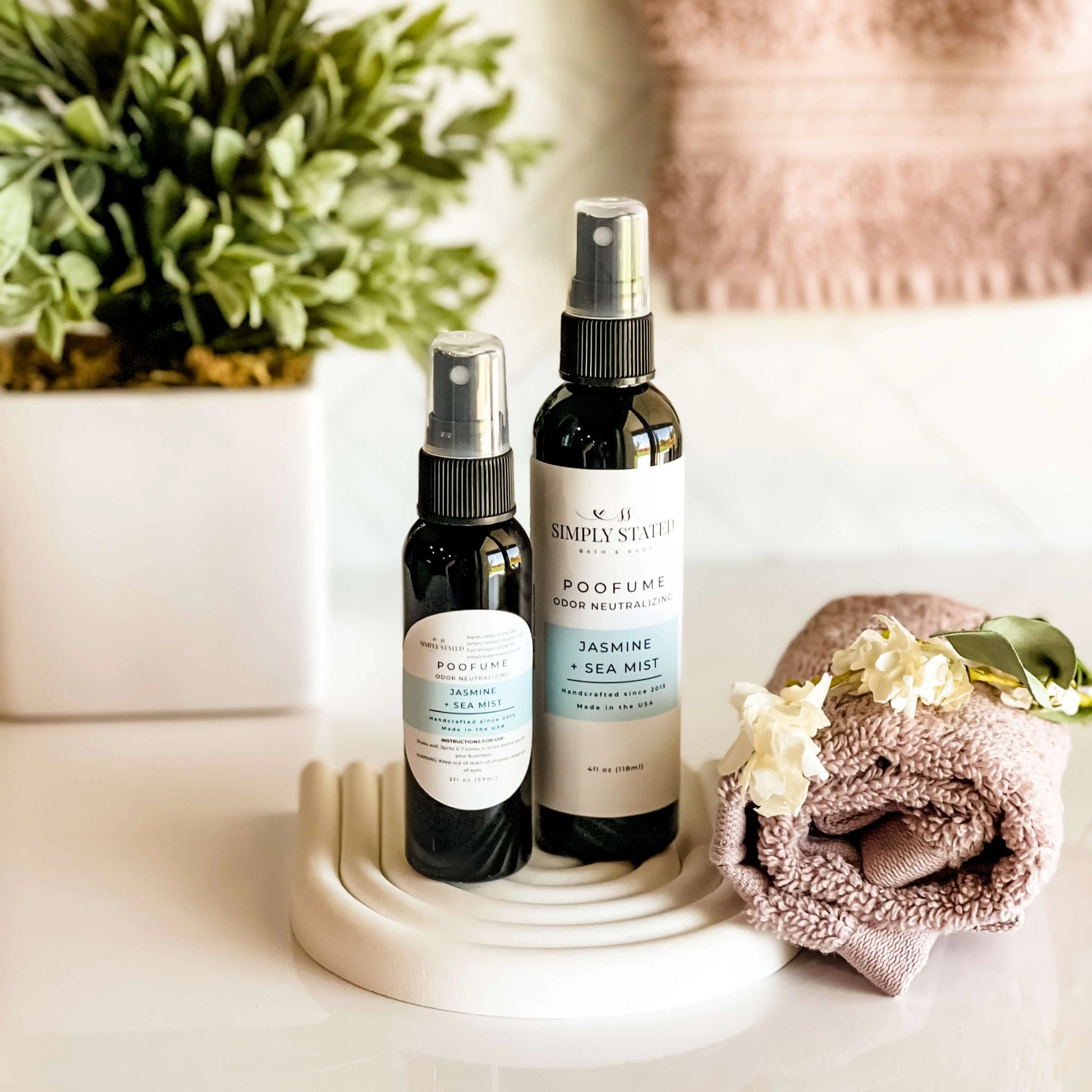 Two spray bottles on a white surface with a plant and towel in the background