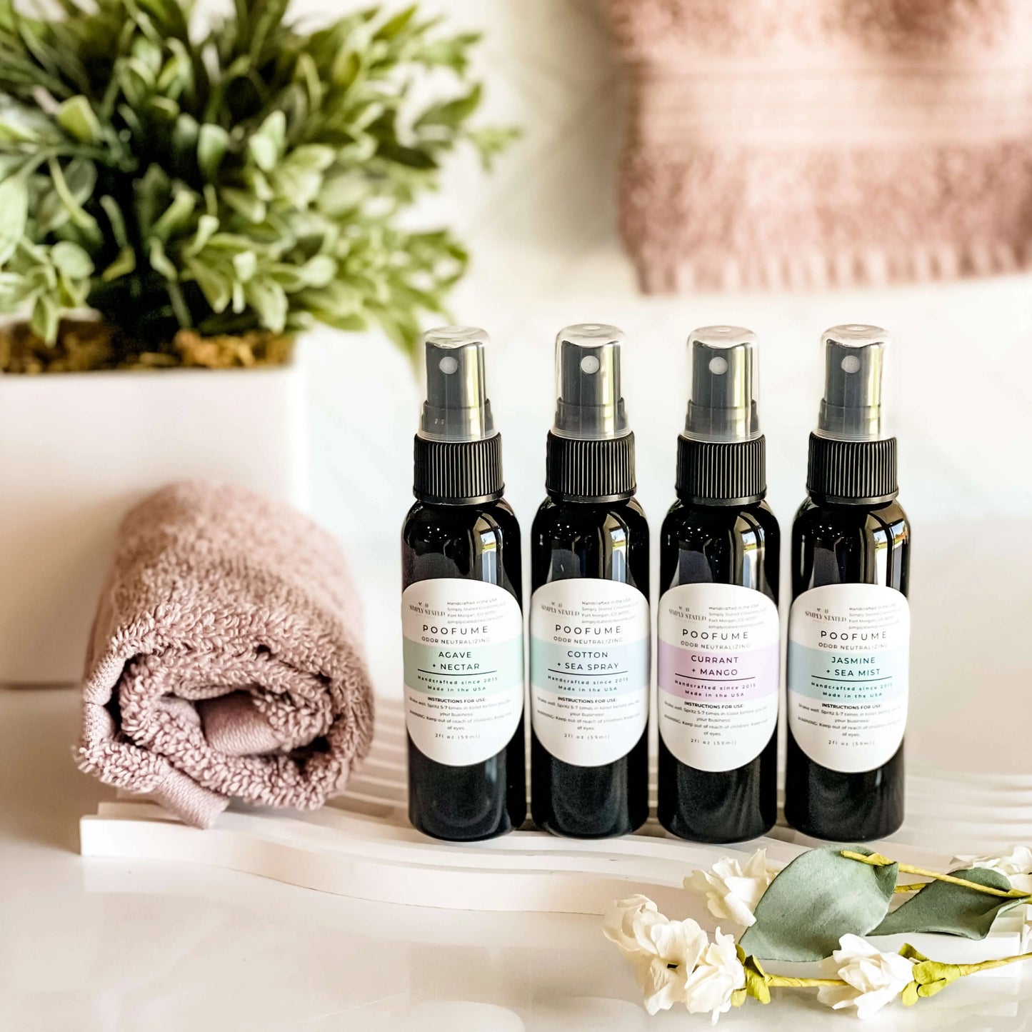 Set of four black spray bottles with labels on a white surface, accompanied by a pink towel and greenery.