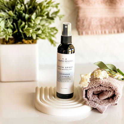 Room spray bottle labeled 'Simply Stated' on a decorative stand with a plant and towel in the background.