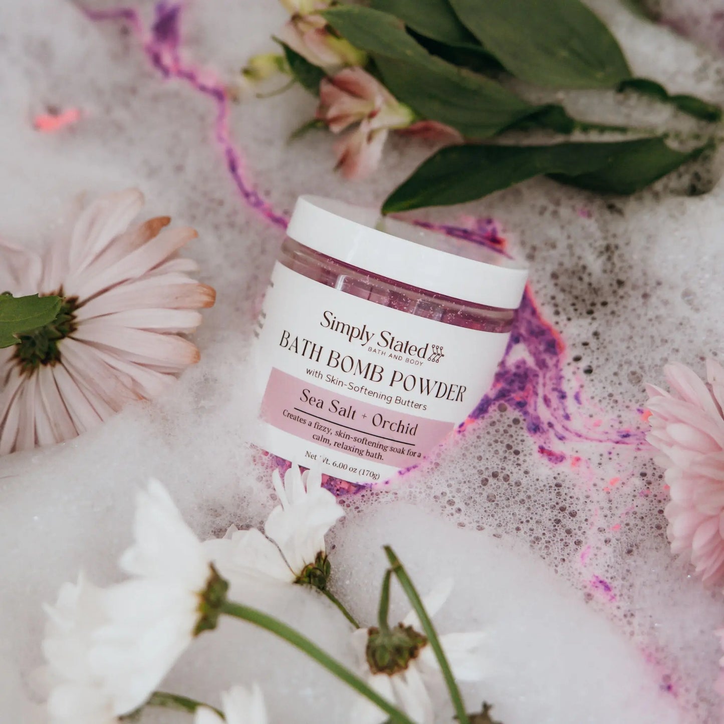 Simply Stated Bath & Body Sea Salt + Orchid Bath Bomb Powder jar lying in pink bubble bath with flowers
