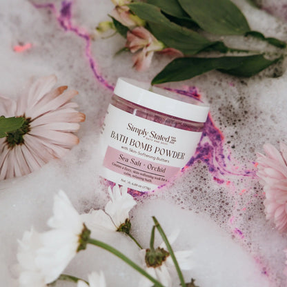 Simply Stated Bath & Body Sea Salt + Orchid Bath Bomb Powder jar lying in pink bubble bath with flowers