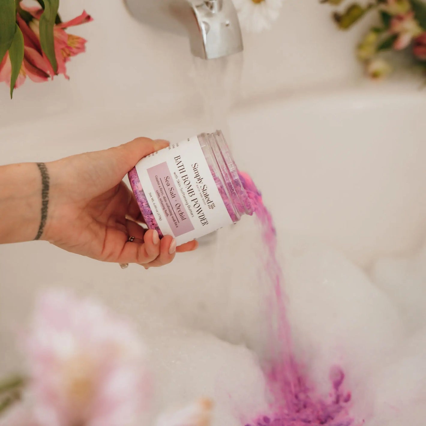 Sea Salt + Orchid Bath Bomb Powder jar being poured into running bathwater with foam and flowers nearby