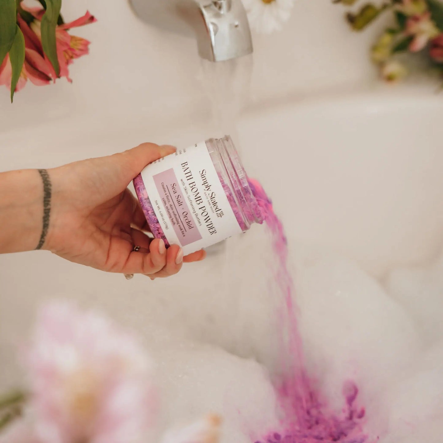 Sea Salt + Orchid Bath Bomb Powder jar being poured into running bathwater with foam and flowers nearby