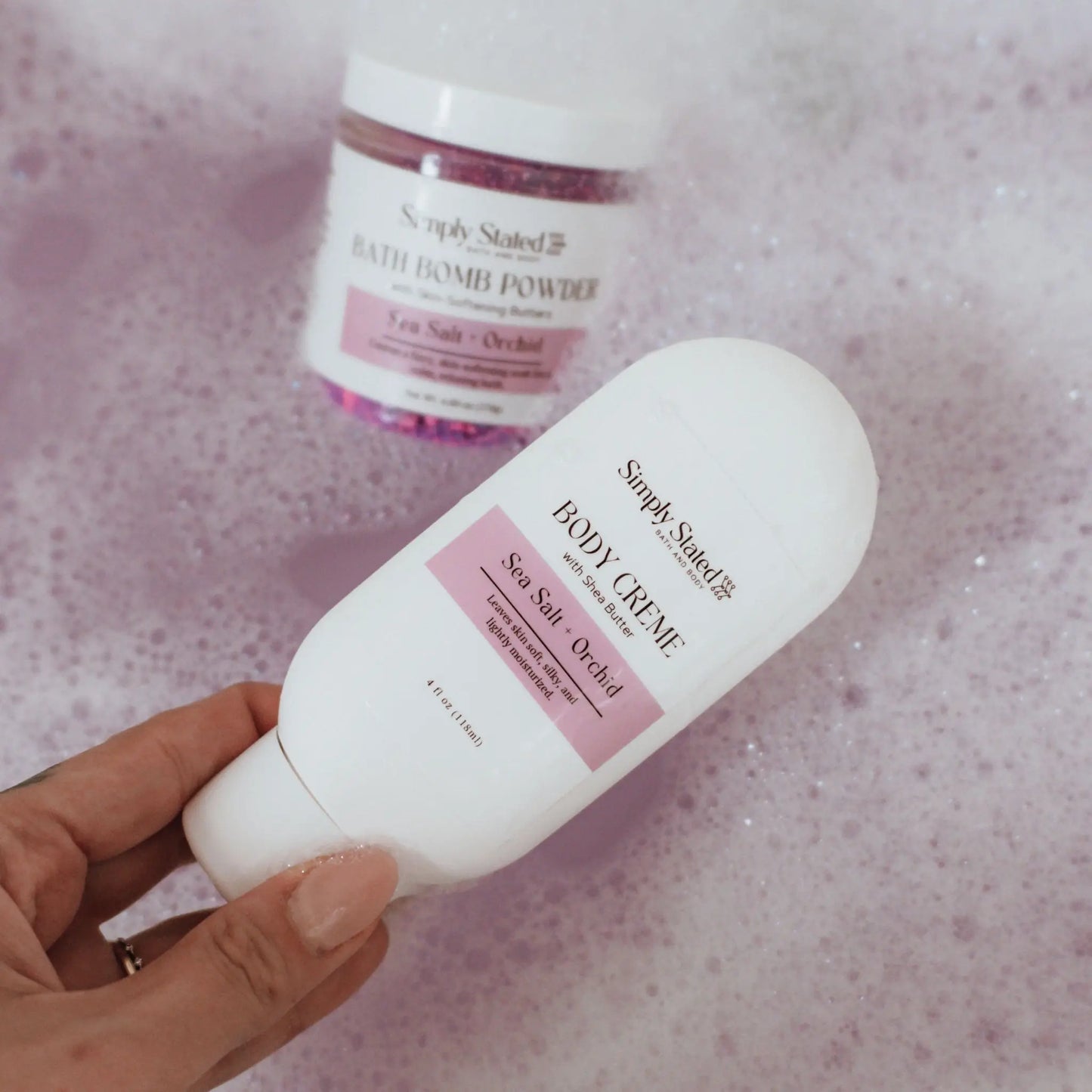 Sea Salt + Orchid Body Creme bottle held over pink bubble bath with Bath Bomb Powder jar in background