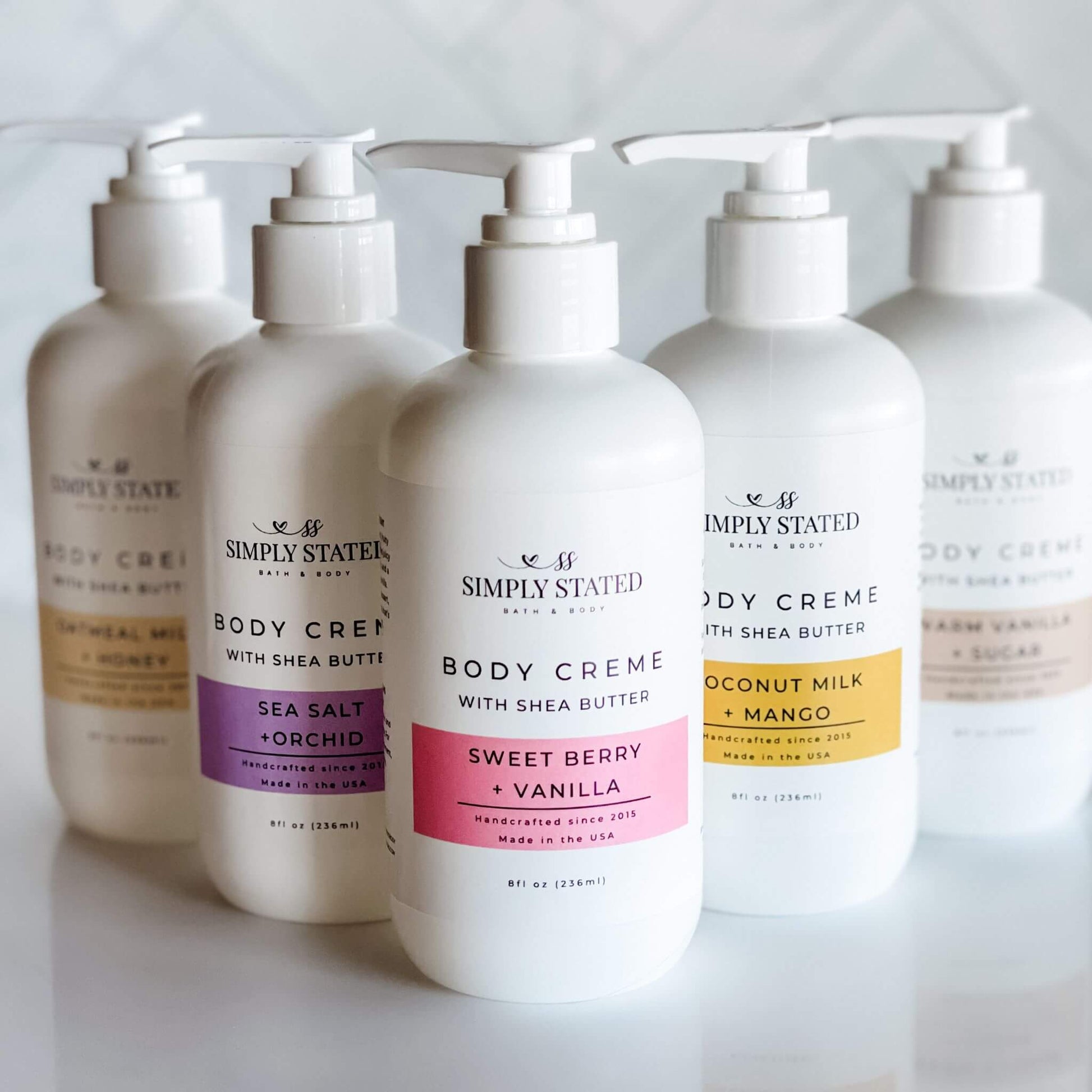 Lineup of Simply Stated Bath & Body 8oz pump bottles for the Signature Body Creme collection, including Coconut Milk + Mango, Oatmeal Milk + Honey, Sea Salt + Orchid, Sweet Berry + Vanilla, and Warm Vanilla + Sugar scents.