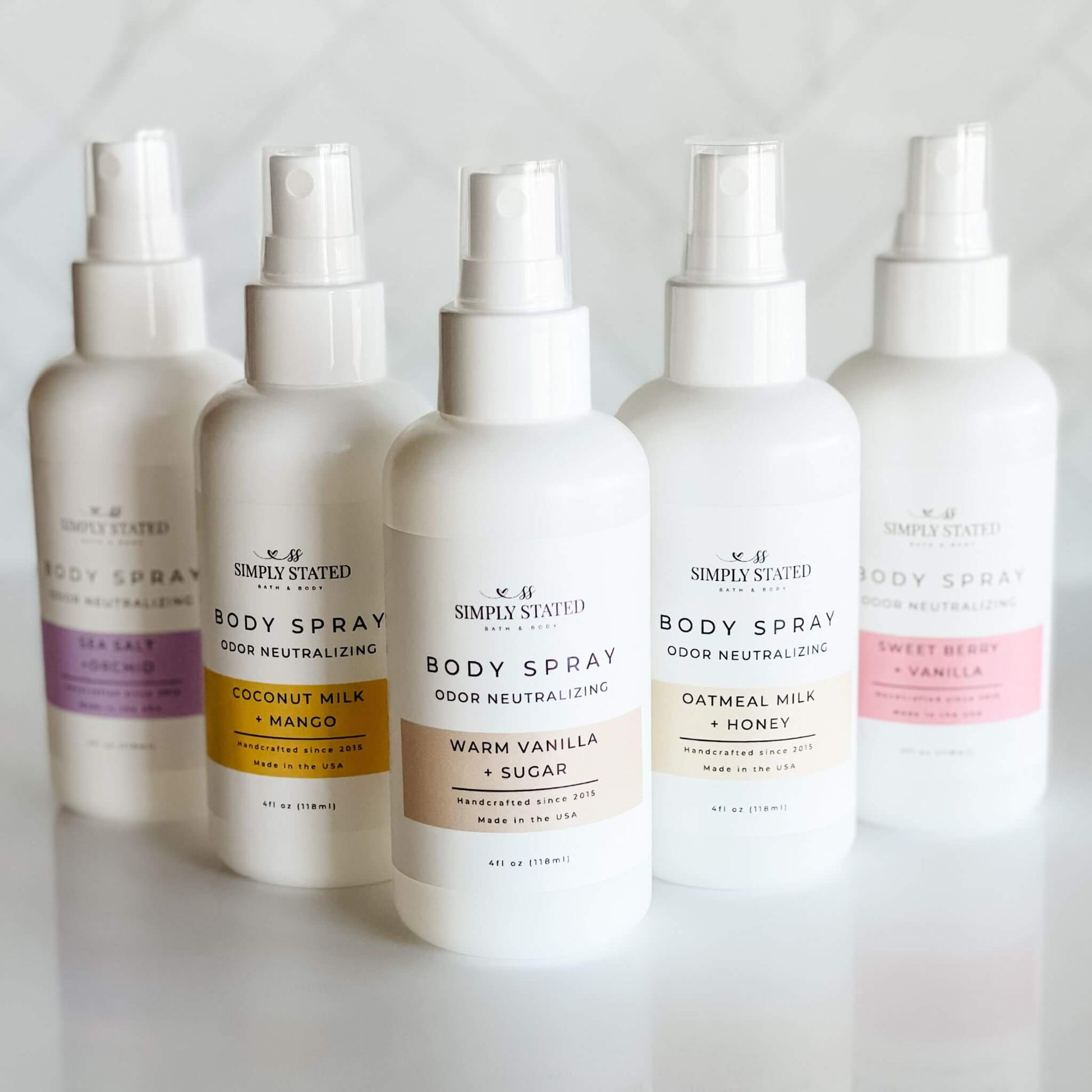 Simply Stated Signature Line Body Spray collection, featuring 4oz white bottles in scents like Coconut Milk + Mango, Oatmeal Milk + Honey, Sea Salt + Orchid, Sweet Berry + Vanilla, and Warm Vanilla + Sugar. Odor-neutralizing formulas in elegant packaging.