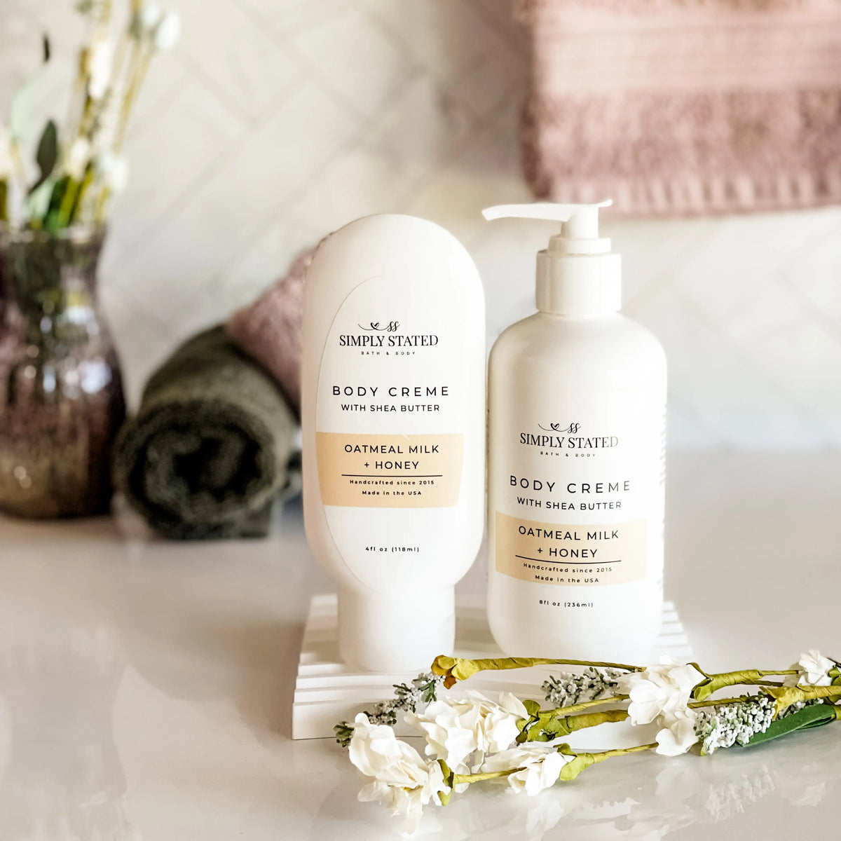 Simply Stated Bath and Body Oatmeal Milk + Honey Body Creme tube and pump bottle on tray with towels and flowers.