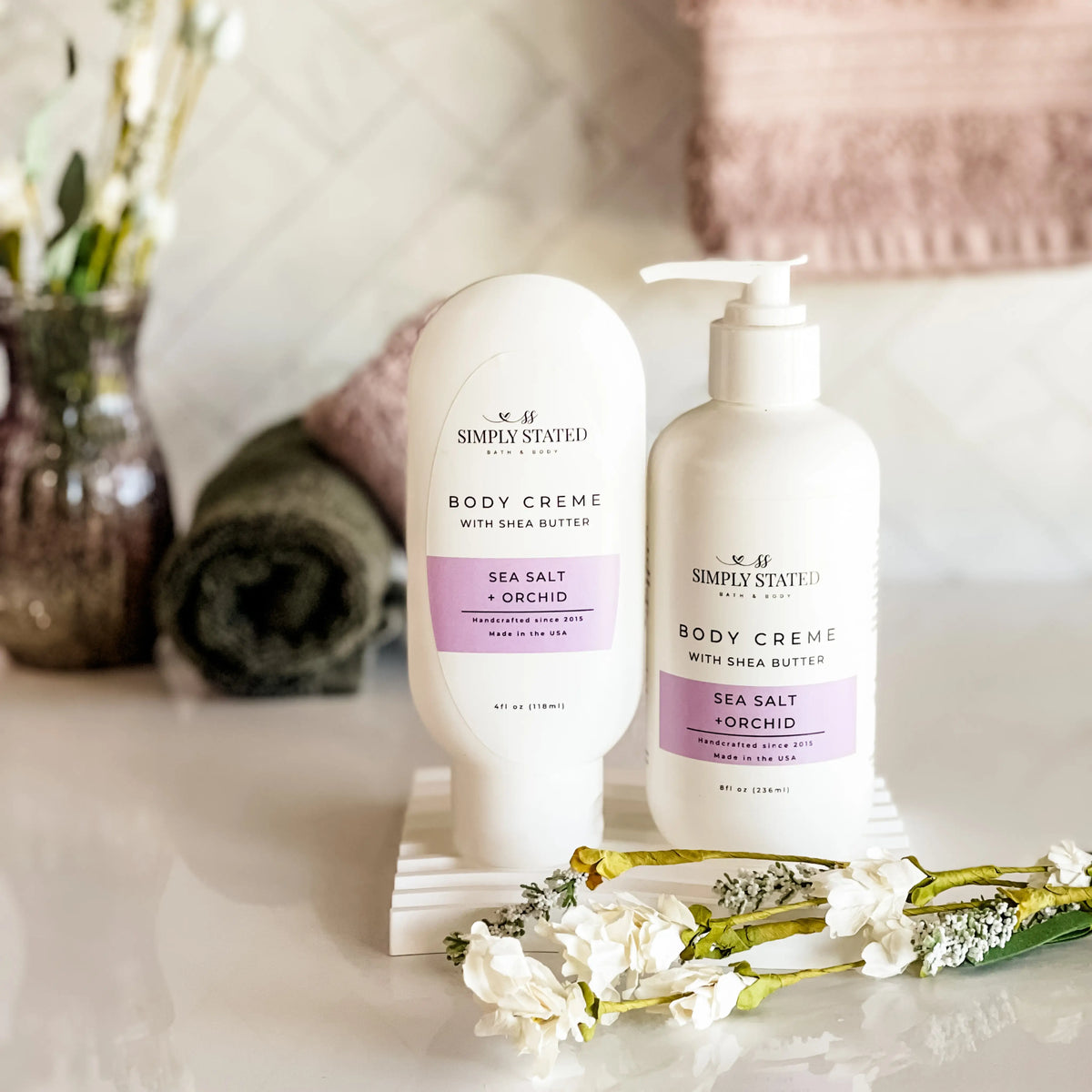 Simply Stated Bath & Body Sea Salt + Orchid Body Creme with Shea Butter bottles, styled with towels and flowers